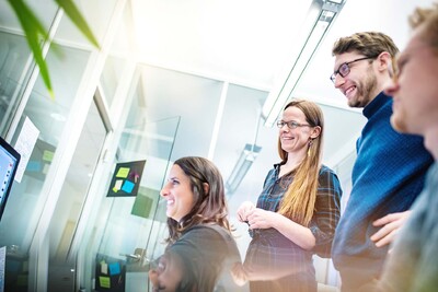 Vier Personen in einem Büro schauen lächelnd auf den Monitor einer der Personen