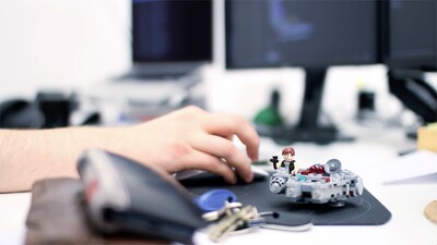 Hand auf Maus auf einem Schreibtisch mit einem Lego-Raumschiff und Bildschirm