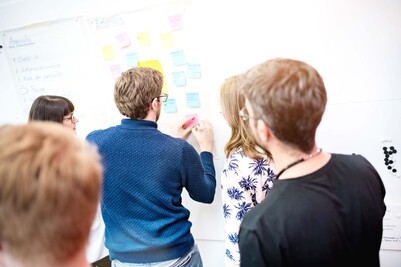 Five people stand in front of a whiteboard with post-its and one of them writes something